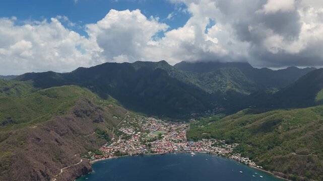 Breathtaking aerial drone footage showcases a vibrant Caribbean coastal town nestled in a valley between dramatic lush green volcanic mountains under partly cloudy blue skies with boats dotting.