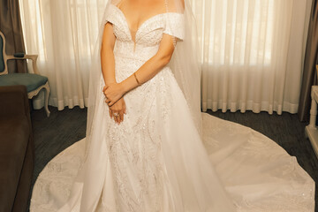 Fototapeta premium Bride in a white dress stands near bright window in a venue before wedding ceremony. bride stands by window wearing white dress with long veil. She is preparing for wedding ceremony