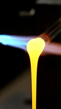 Molten yellow glass drips down from a blowpipe while being heated by a blue and orange torch flame in a dark workshop setting for glassblowing artistry and craftsmanship