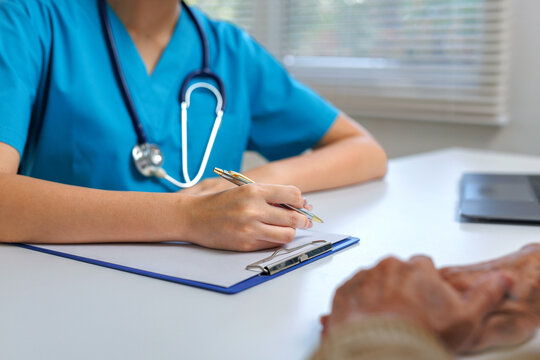 Doctor in blue scrubs writing on a clipboard during a healthcare consultation with a senior patient, providing medical care