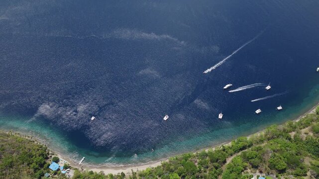 Stunning aerial drone view of a tropical bay in Saint Lucia with multiple white boats anchored in deep blue waters, one boat creating wake trails, lush green forested coastline and sandy beach below.