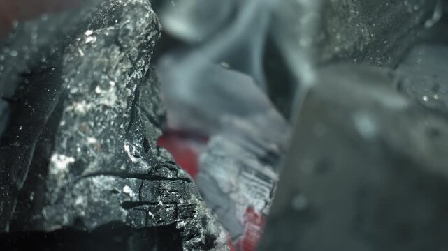 Macro closeup smoldering coal with smoke, rising pale plume and drifting ash flakes, glowing red embers peeking through cracked charcoal, soft cinematic motion and cooling atmosphere