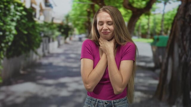 Young woman clutching her throat with both hands on a tree lined street, eyes closed and grimacing; panic.