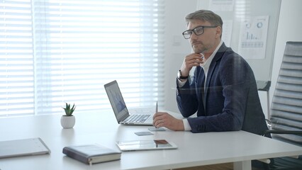 Fototapeta premium Businessman concentrating on a laptop screen displaying charts, thinking deeply while working at a modern office desk, reflecting on data, and strategizing future business development