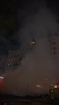 Two firefighter in protective gear amid thick white smoke on a city street at night, orange flame and illuminated building visible, crowd in background during fire festival