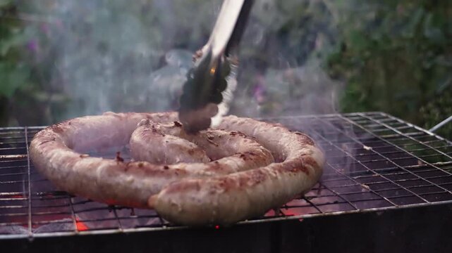 Macro closeup sausage flipped by tongs on charcoal grill, smoking coals and sear marks forming, sizzling juices and charred texture, lively outdoor cook gesture, garden patio setting