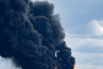 Black plumes of smoke and towering flames from a massive industrial fire over a cloudy sky,...