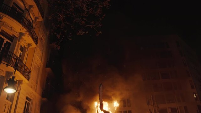 Large fire burning on narrow city street at night during Fallas festival in Valencia, spark and ember flying past ornate illuminated building with iron balcony