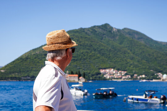 Mature man with straw hat walks seaside and looking at boats on sea in Perast, Montenegro