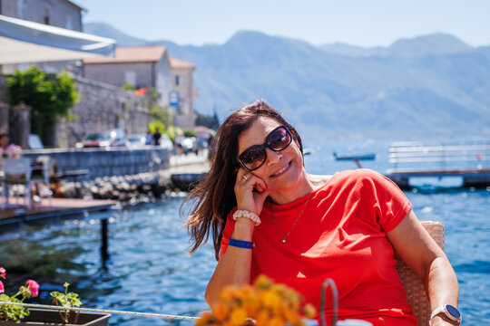 Happy smiling mature woman sitting in outdoor restaurant at sea bay in Kotor city in Montenegro. Enjoyment of summer holiday
