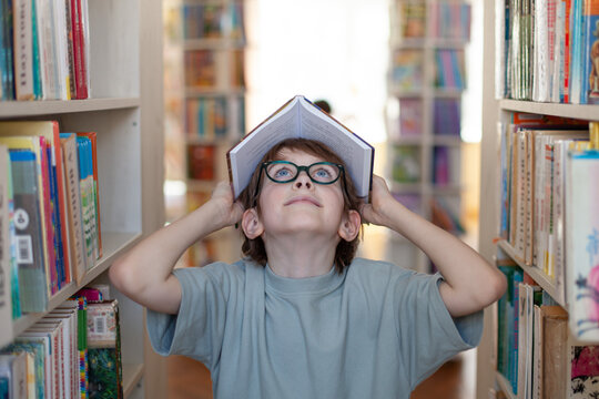 Child stands between library shelves holding open book over head. Concept of reading, childhood curiosity and playful learning.