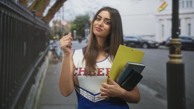 Young woman cheerleader in blue and white uniform holding a yellow clipboard and books, beckons with finger and smiles on a street beside iron railings; confidence youth.