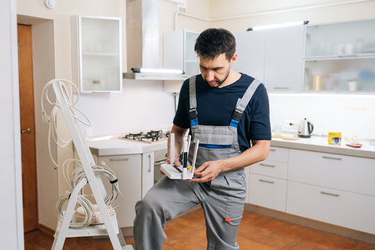 Focused technician in uniform installs network router for home internet. Man holding device in modern kitchen with stepladder and cables nearby. Professional service and connectivity concept.