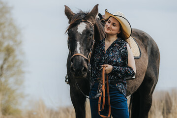 Obraz premium Young woman in floral shirt and cowboy hat stands beside brown horse holding reins in a grassy field with trees and cloudy sky in the background