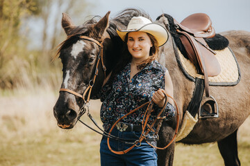 Young woman in cowboy hat stands beside brown horse with saddle, holding reins in a sunny outdoor setting with grass and trees in the background © Emvats