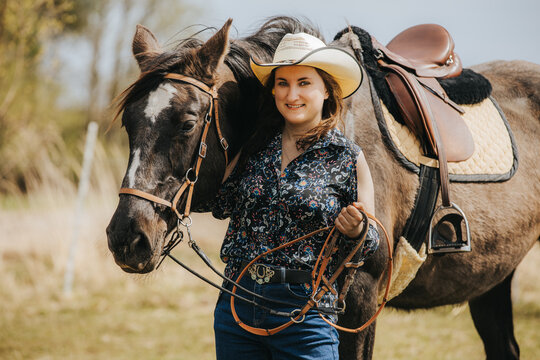 Young woman in cowboy hat stands beside brown horse with saddle, holding reins in a sunny outdoor setting with grass and trees in the background