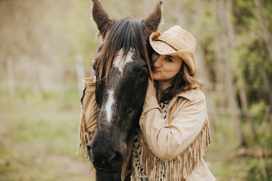 Young woman in a straw hat and fringed jacket poses affectionately with a black horse in a natural outdoor setting surrounded by greenery