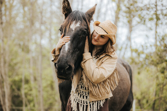Young woman in a fringed jacket and straw hat gently embraces a black horse outdoors, surrounded by trees and natural greenery in a serene rural setting