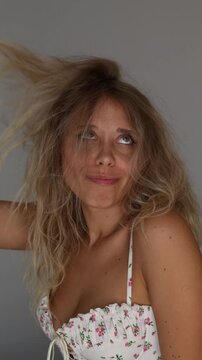 Young blonde woman lifting messy frizzy hair and making funny faces. Playful bad hair day concept, self irony, volume, dry porous bleached hair, carefree mood on neutral background