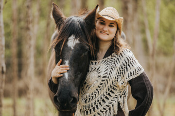 Obraz premium Young woman wearing a straw hat and crochet poncho stands beside a black horse in a forested area with trees and greenery in the background
