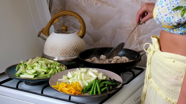 Traditional village kitchen scene with a woman preparing homemade food on an old gas stove, mixing meat with zucchini, carrots, pattypan squash and green beans in a worn black frying pan.