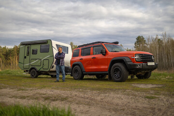 Man stands with arms crossed between a green camper and an orange SUV in nature © WoodHunt