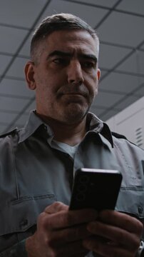 Serious Security Officer With Prominent Arm Tattoos Stands by Grey Metal Locker, Concentratedly Typing Message or Email Response on Smartphone After Finishing Airport Security Shift. Vertical Shot.