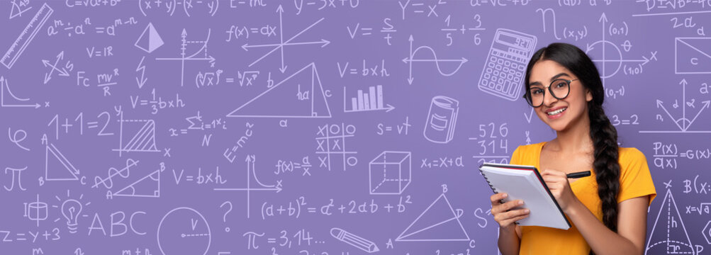 A student stands in a classroom setting with a purple collage of math and science drawings behind her. She is holding a notepad and writing while looking at the board. It is daytime.