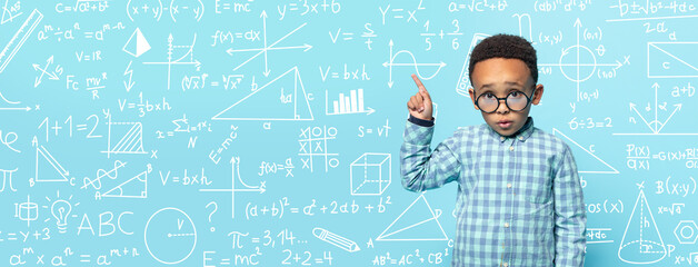 A child stands in front of a blue wall filled with math formulas and graphs. The child raises a finger, indicating a point of interest. © Prostock-studio