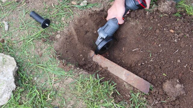 Worker using a cordless angle grinder to cut a rusty metal pipe buried in the ground, creating bright sparks and removing an obstacle during outdoor earthworks