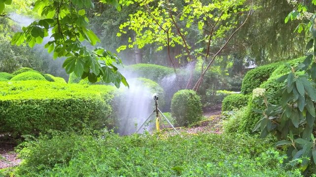 Automatic tripod water sprinkler spraying mist over lush green garden bushes and trees