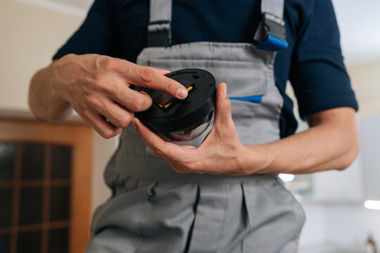Close-up of male technician in uniform holding dome surveillance camera. Professional worker installs smart home security system for safety and protection. Focus on hands and equipment.