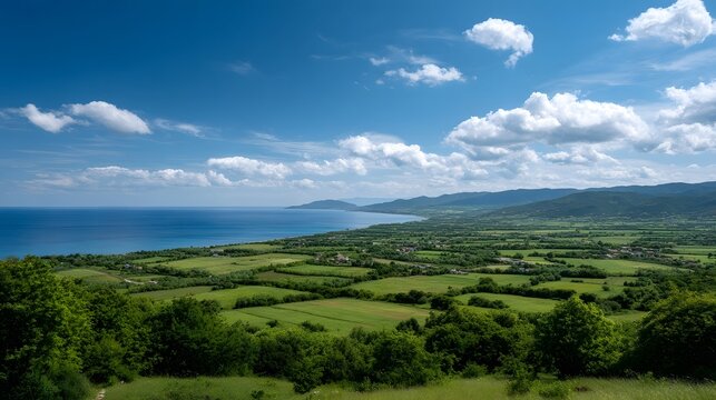 Panoramic view of lush green fields and scattered settlements leading down to a vast bright blue sea under a dynamic sky