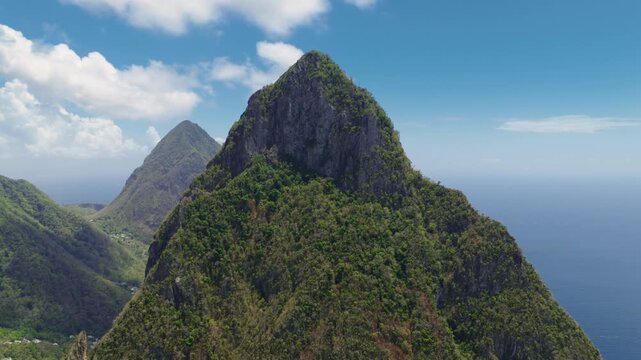 Breathtaking aerial drone footage captures majestic Pitons of Saint Lucia rising dramatically from lush tropical rainforest, with rocky volcanic peaks against bright blue sky dotted with white clouds.