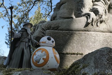 Naklejka premium Action figure of fantasy wizard Gandalf known from novels Hobit and Lord Of The Rings, following spherical Star Wars astromech droid BB8 on mossy path next to huge stone lion statue.