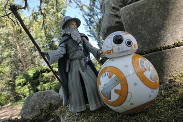 Naklejka premium Action figure of fantasy wizard Gandalf known from novels Hobit and Lord Of The Rings, following spherical Star Wars astromech droid BB8 on mossy path next to huge stone lion statue. Park background. 
