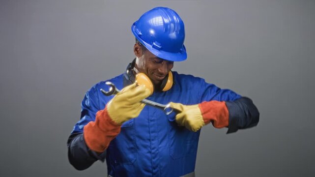 Man in blue hardhat and work coveralls inspects a wrench with yellow earmuffs and gloved hands in studio; diligence duty.