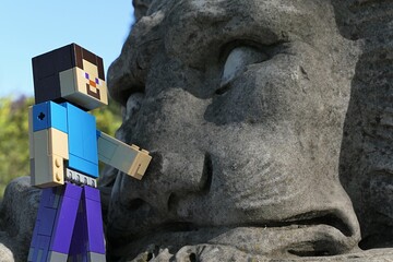 Obraz premium Action figure of main Minecraft Character Steve standing next to a face of giant lion statue in park, spring blue skies in background. 