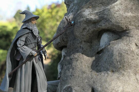 Detail of action figure of wizard Gandalf from Tolkien novels Hobit and Lord Of The Rings touching a large stone head of a lion with his staff. Spring nature in background. 