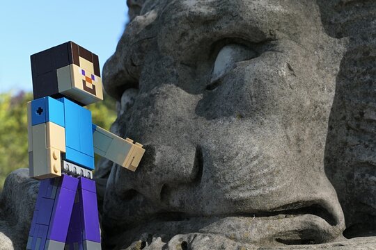 Action figure of main Minecraft Character Steve standing next to a face of giant lion statue in park, spring blue skies in background. 