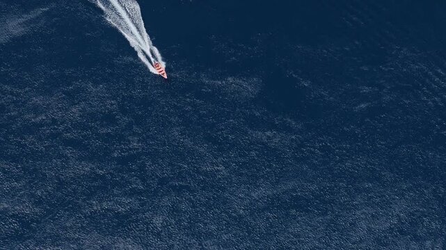 Aerial drone footage captures a red and white speedboat racing across vast deep blue ocean waters leaving a dramatic white wake trail behind. A catamaran appears in the distance.