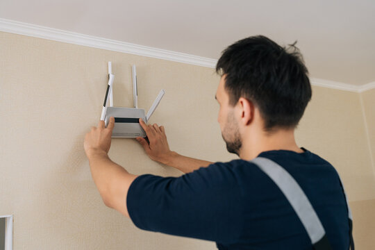 Male technician in uniform mounts wireless router on beige wall. Handyman setting up home network for internet access. Technology, connectivity and installation service concept.