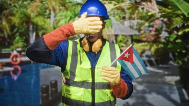 Man construction worker holding cuban flag and covering eyes with gloved hand near pool building and hardhat visible; patriotism pride.