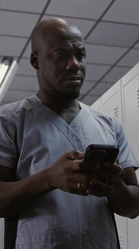 African American Male Doctor Looking Phone With Shocked Expression, Appearing Confused and Saddened by Urgent News in Professional Hospital Setting. Concept of Human Side of Medicine. Vertical Shot.