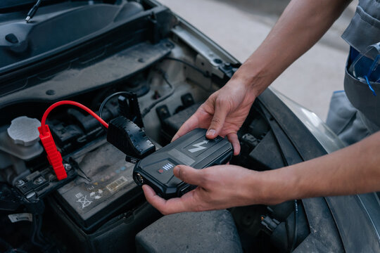 Close-up of mechanic hands connecting red clamp of portable jump starter to dead car battery. Automotive service concept for vehicle breakdown assistance and roadside repair.