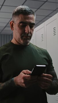 Focused Man in Camouflage in Locker Room, Concentratedly Typing Message to Family on Smartphone Before Departing For Mission. Personal Moment in Military Life in Base Dressing Room. Vertical Shot.