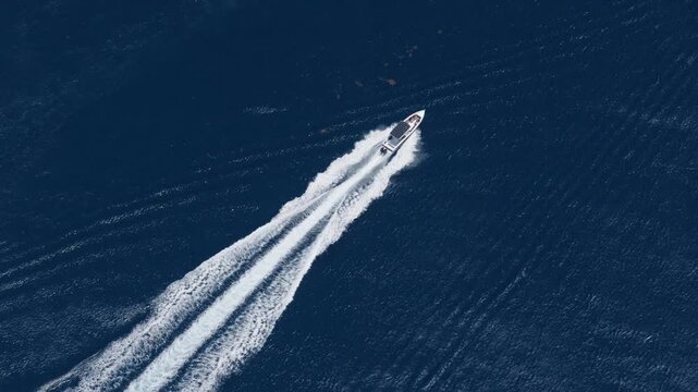 Aerial drone view of a sleek white luxury speedboat racing diagonally across deep navy blue ocean waters, leaving a long dramatic white foamy wake trail as a smaller blue boat enters frame.