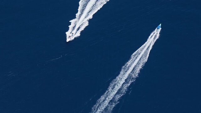 Aerial view of two motorboats speeding across dark blue ocean water leaving dramatic white foam wake trails behind them. Boats travel in parallel diagonal paths across open sea.