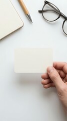 Obraz premium Hand holding blank white business card with notebook, pen and glasses on clean desk workspace for corporate branding and professional identity design.