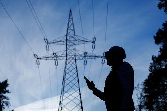 Silhouette of a male power engineer with a phone in his hand stands against the background of a power line and a blue sky.